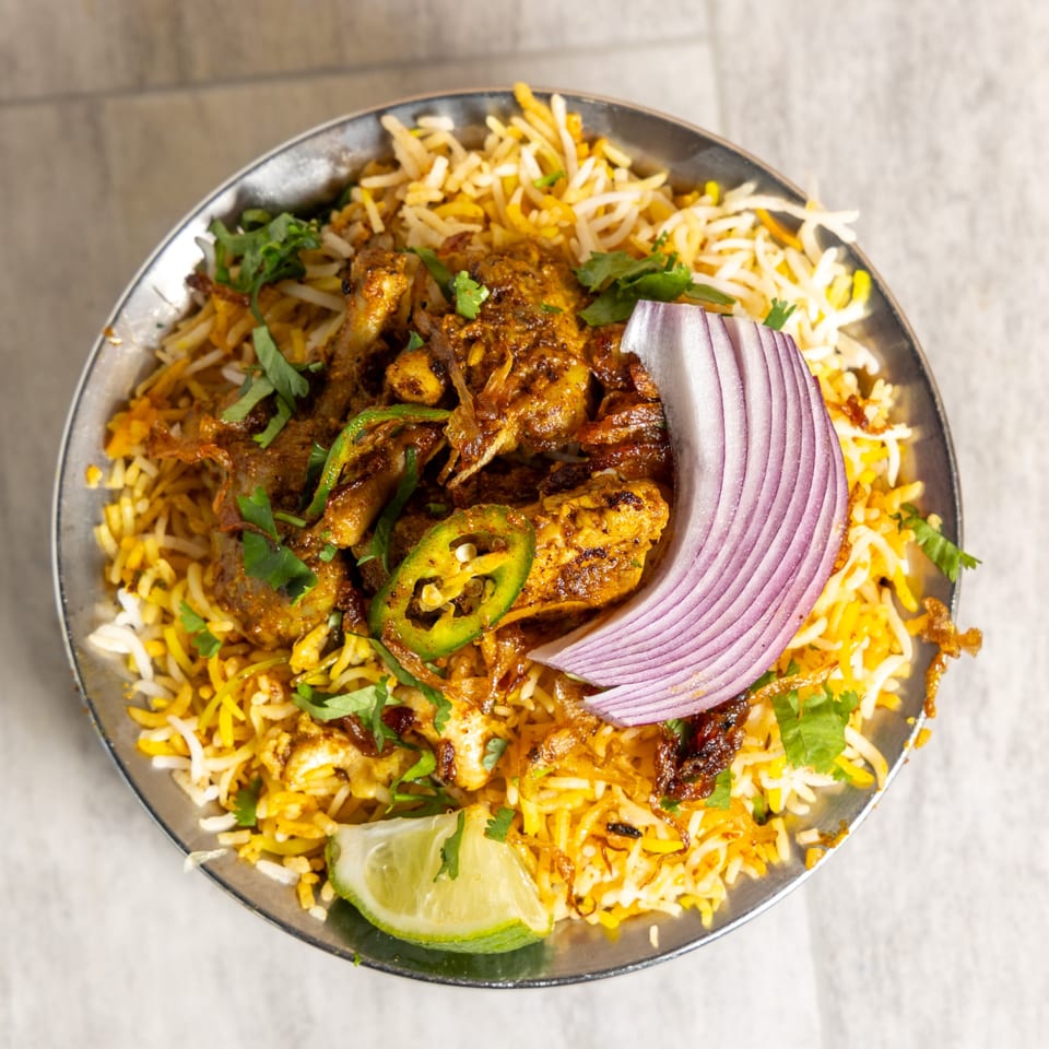 Chicken biryani with rice, red onion, green chilies, cilantro, and a lime wedge in a metal bowl.