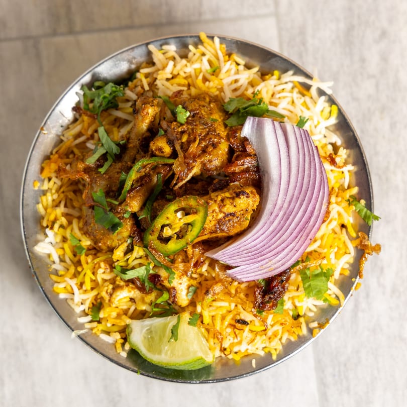 Chicken biryani with rice, red onion, green chilies, cilantro, and a lime wedge in a metal bowl.