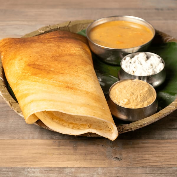 Golden brown dosa served with three small bowls of chutney and sambar on a banana leaf.