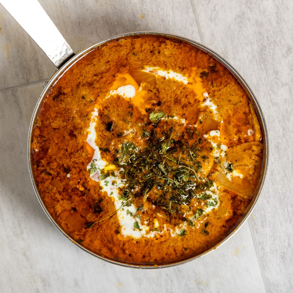 A close-up of a pan filled with a rich orange curry, garnished with cream swirls and fresh herbs.