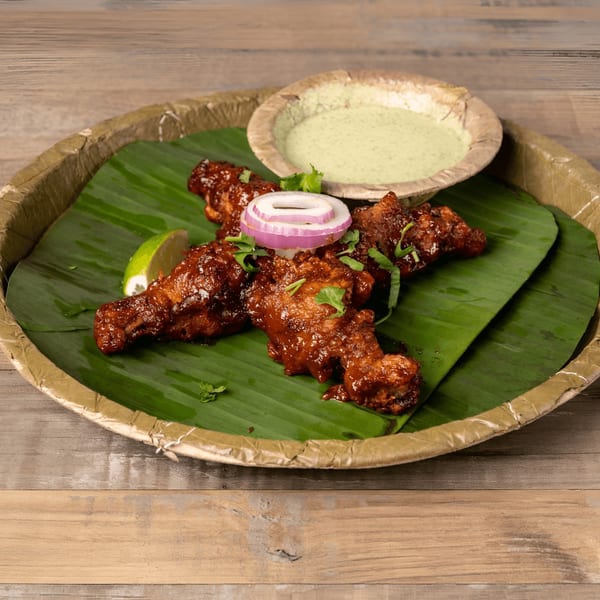 Spicy chicken drumsticks on a banana leaf with lime, onion, cilantro, and green dipping sauce.