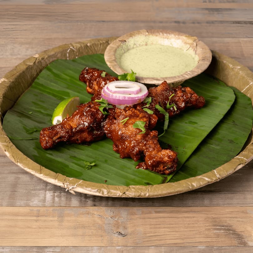 Spicy chicken drumsticks on a banana leaf with lime, onion, cilantro, and green dipping sauce.